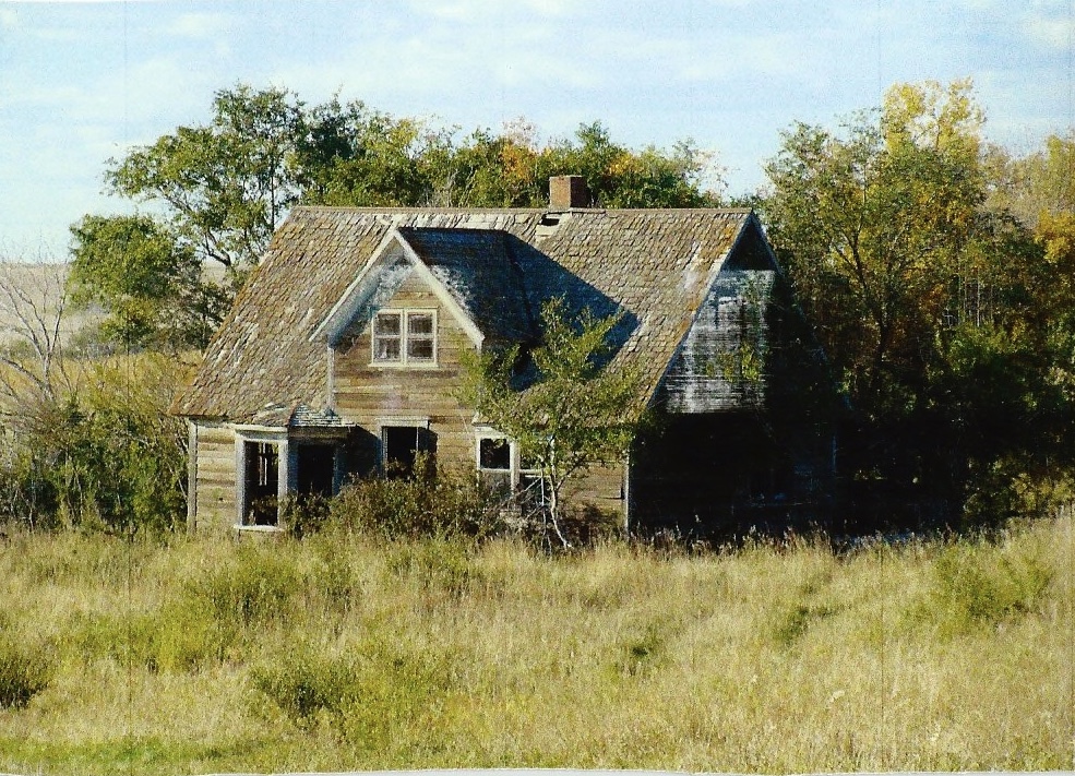 A weathered prairie house with projecting window bays, surrounded by tall grass and trees.
