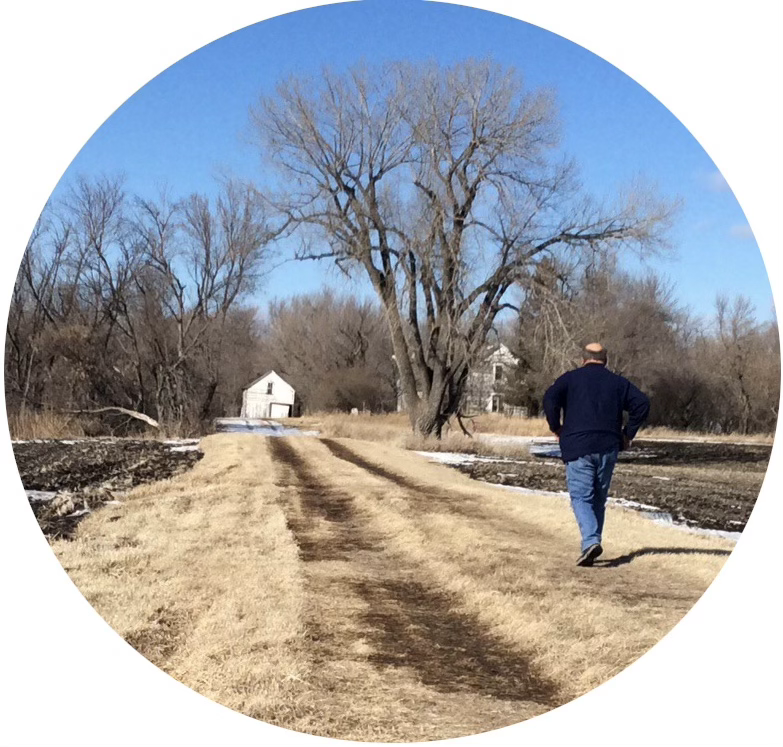 A man walking along a grassy farm lane toward bare trees and old buildings.
