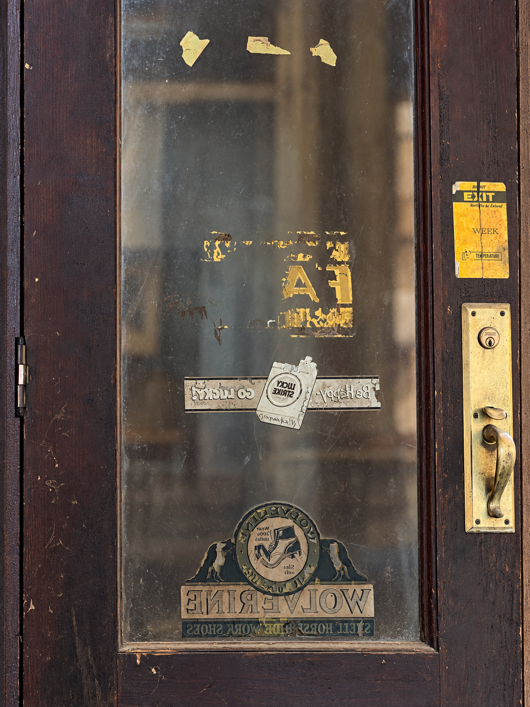The store door seen from inside, with its worn brass handle and surviving reversed Lucky Strike and Wolverine decals.